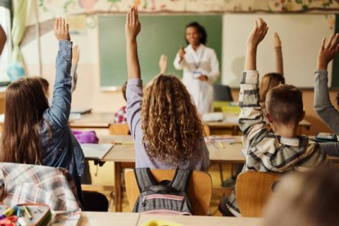 Room of primary kids back sitting with hands up, teacher at the front