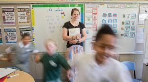 A young teacher stands against a whiteboard at the front of her classroom with happy kids running in a circle.